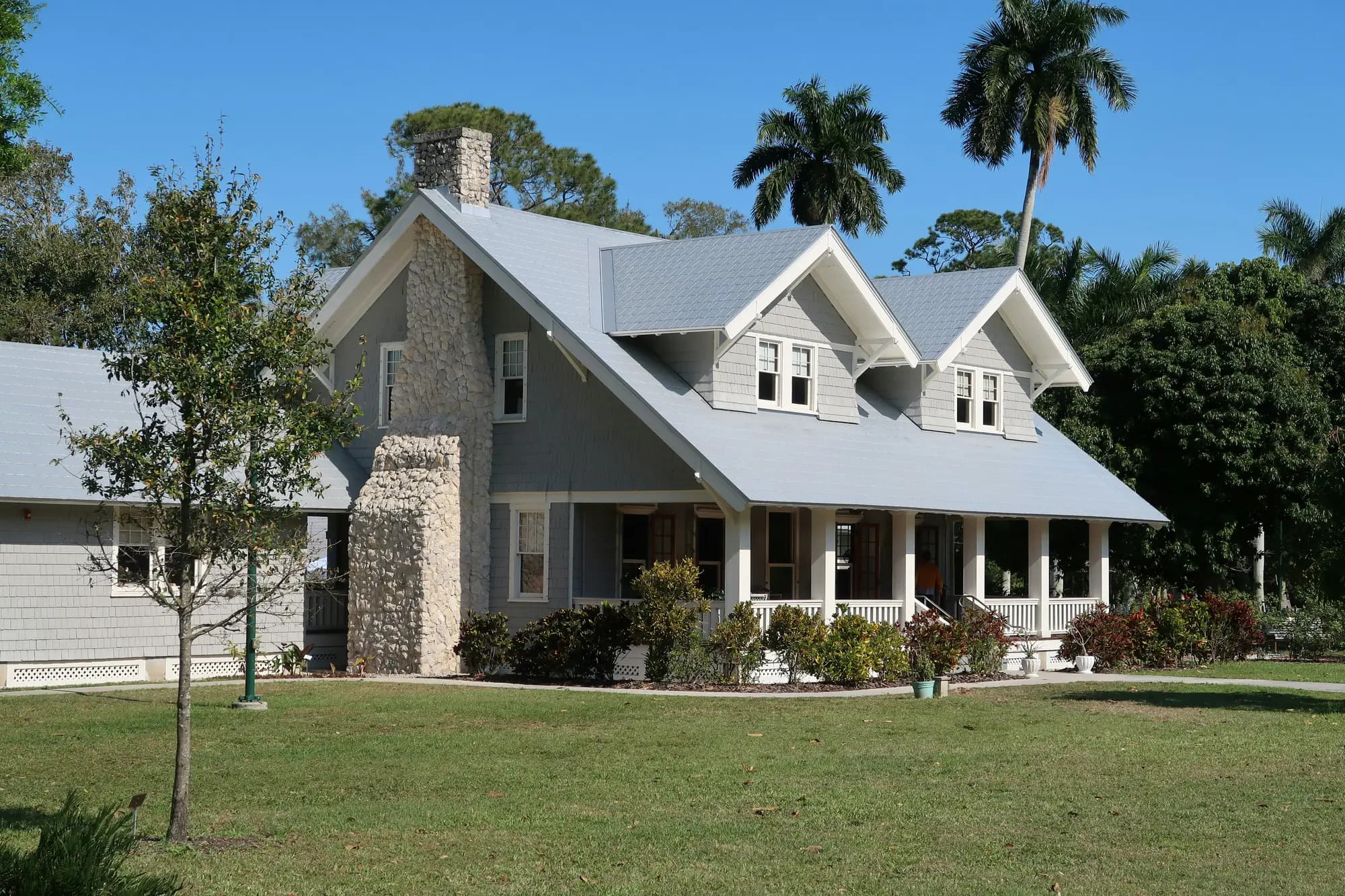 Gray-and-white craftsman home with a wraparound porch, palm trees and clear blue sky