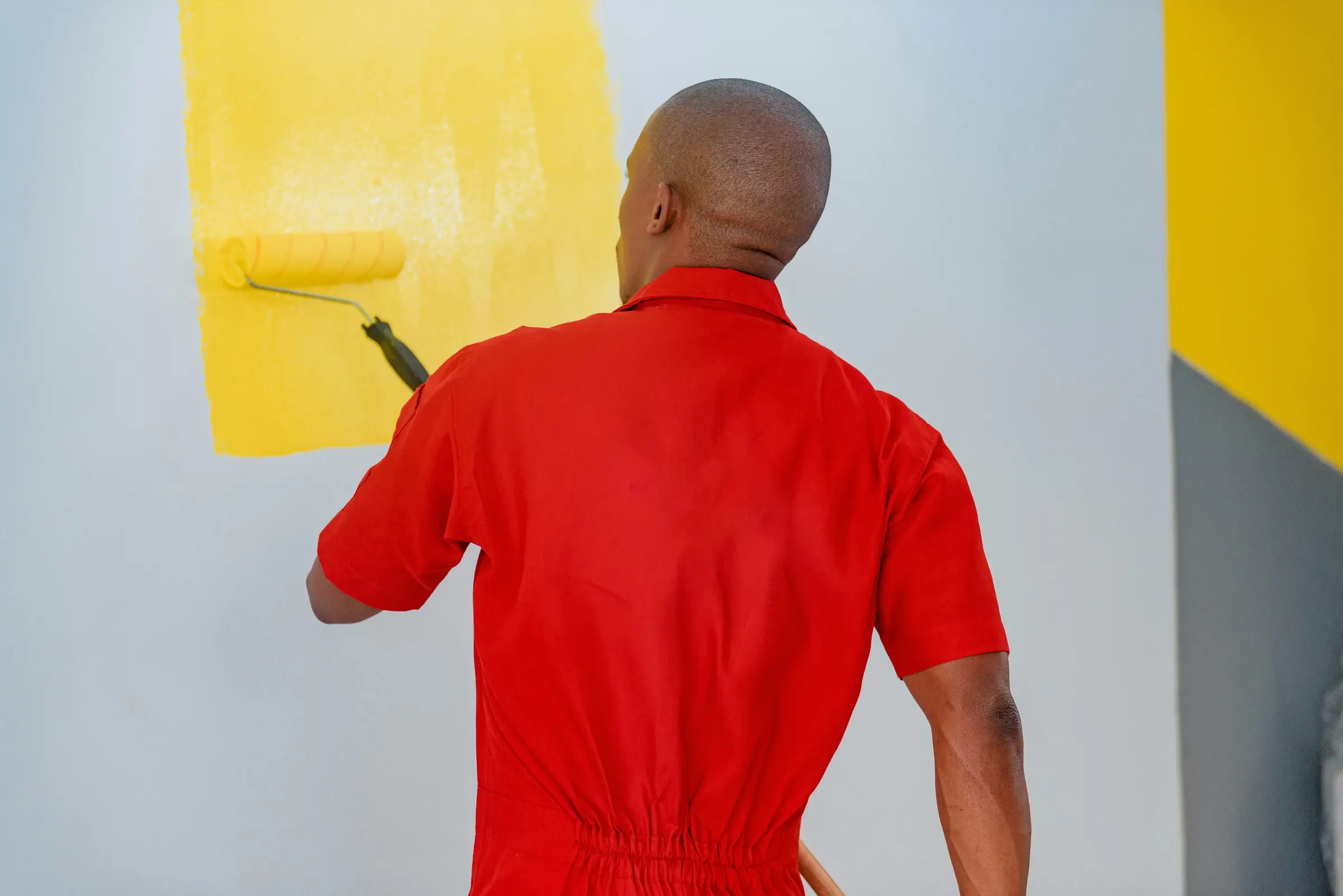 Painter in red coveralls, seen from behind, rolling yellow paint onto a wall